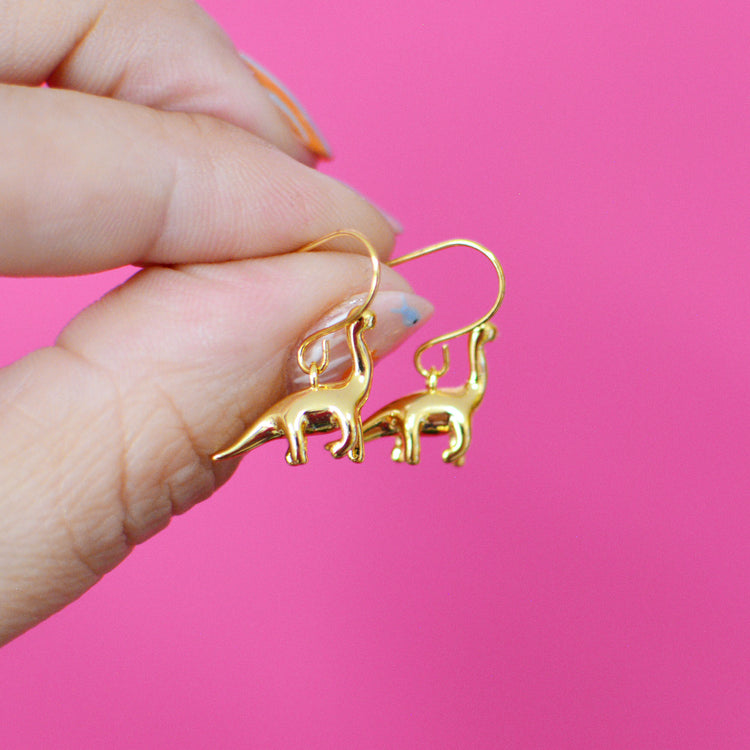 Gold plated brontosaurus shaped earrings with hooks, held by a hand against a pink background