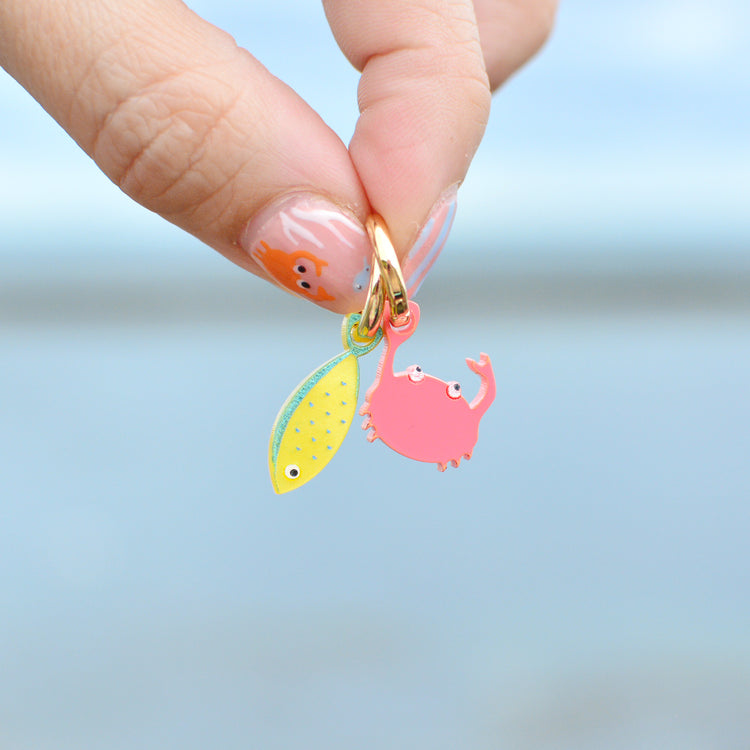 A close-up of a hand holding a gold-tone huggie hoop earring with two charms: a yellow fish with a blue stripe and a pink crab, against a blurred blue background.