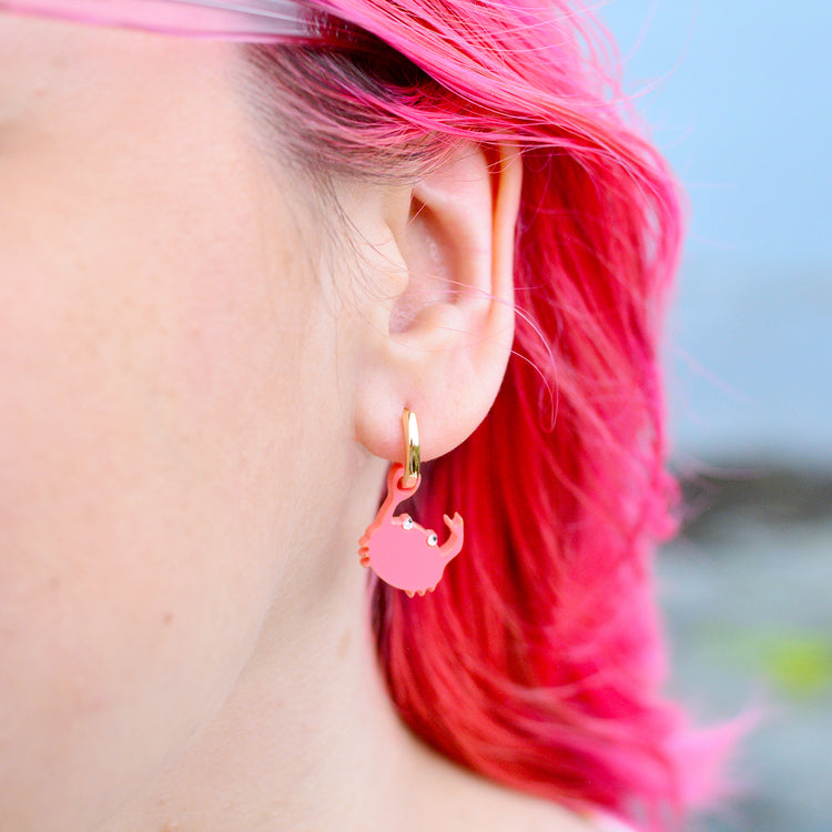 A close-up of a person's ear wearing a small pink crab-shaped dangle earring with a gold hoop against a blurred outdoor background.