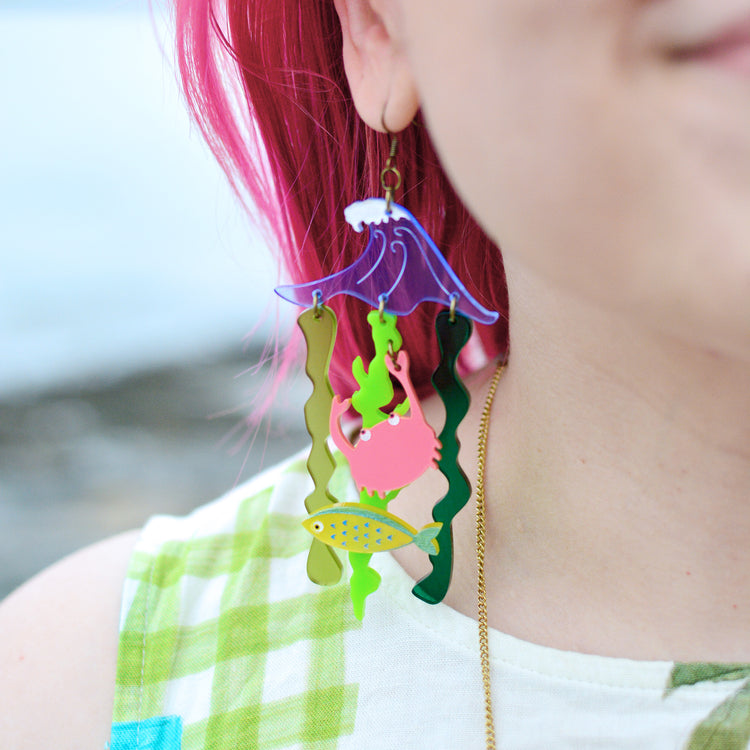 A close up of a woman's ear with her pink hair showing in the background against a blurry picture of the sea. She is wearing a statement earring of a blue wave with green seaweed hanging below. There is also a yellow fish and a coral pink crab attached to the seaweed.