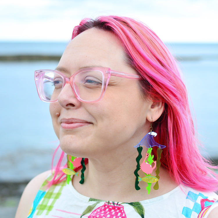 A woman with pink glasses and pink hair stands in front of the sea wearing two statement earrings that feature a blue wave shape with green seaweed ribbons hanging below. The earrings also feature a yellow fish and a coral pink crab hanging from the seaweed.