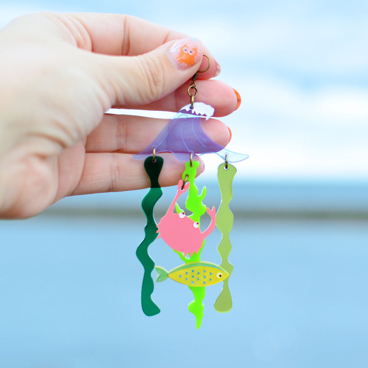 A very large earring being held up in front of the sea by a woman's hand. The earring has a sea wave shape in blue with green wiggly seaweed pieces hanging below. There is also a yellow fish and a coral pink crab hanging from the seaweed.