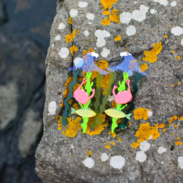Very large earrings featuring a blue wave shape with green seaweed shapes dangling from it. There is also a yellow fish and a coral pink crab hanging from the seaweed. The background is a rock near the sea covered in moss.