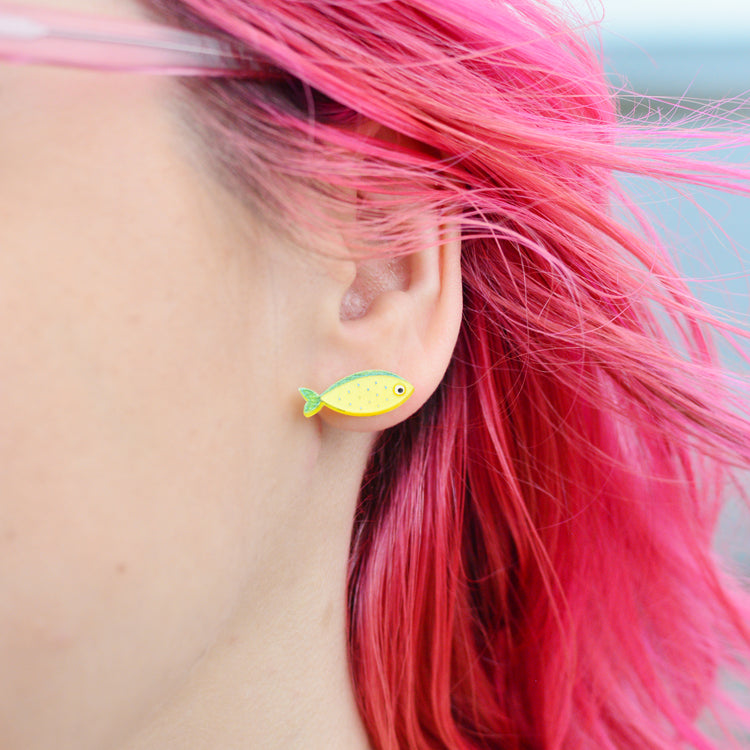 A woman's ear is shown close up against the backdrop of her pink hair and the sea. She wears an earring that is a yellow fish shape with blue details and a big white eye with black centre.
