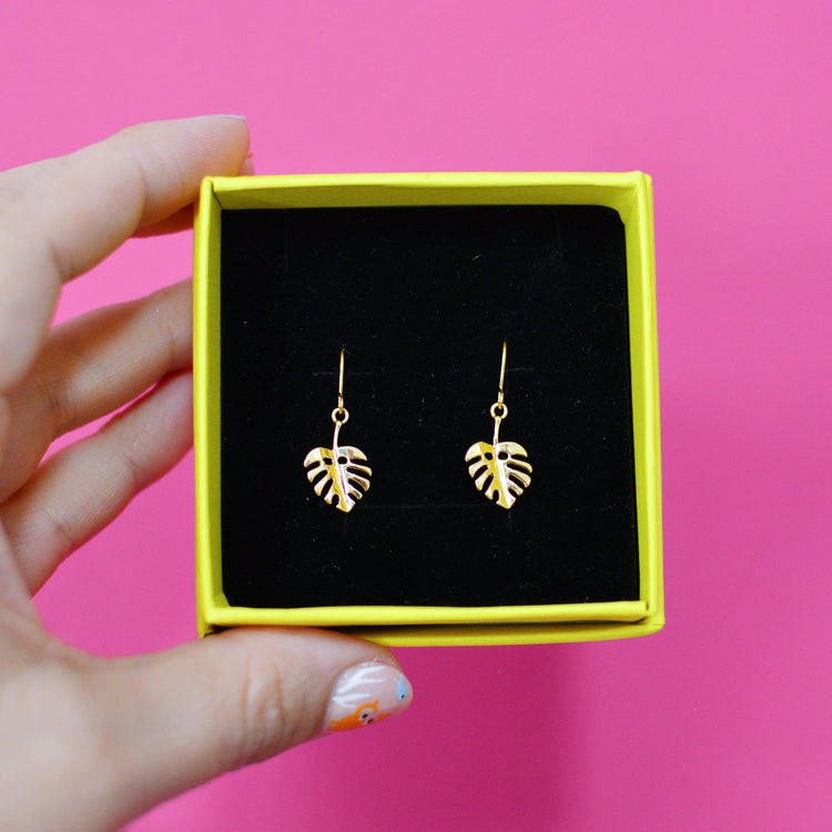 Gold plated monstera leaf earrings in a yellow box with black velvet padding, held by a hand against a pink background