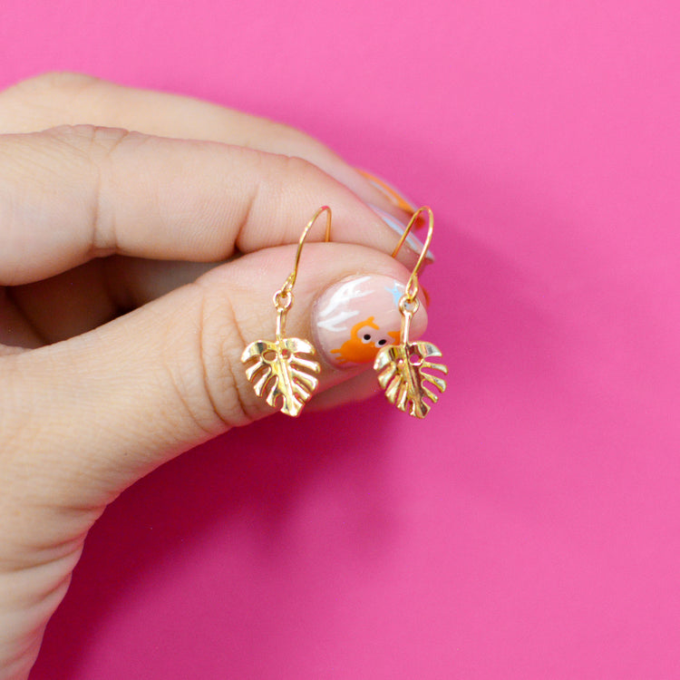 Gold leaf earrings held by a hand against a pink background