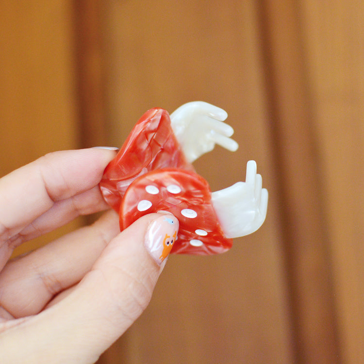 Hand holding a red and white mushroom design hair claw clip against a wooden background. The claw is held open to show the teeth that will hold your hair together.