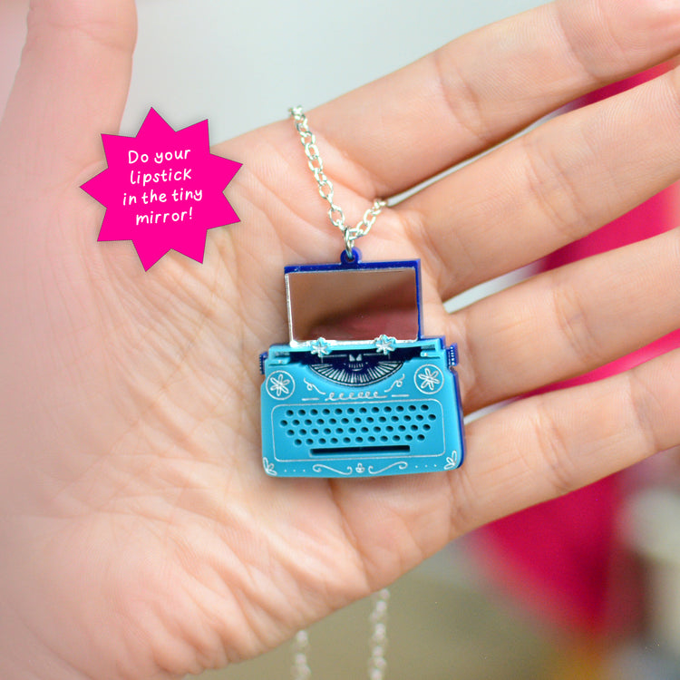 A hand holding a miniature blue typewriter pendant necklace with a tiny mirror where the paper would be inserted, suspended from a silver chain. A pink starburst graphic in the top left reads 'Do your lipstick in the tiny mirror!'.