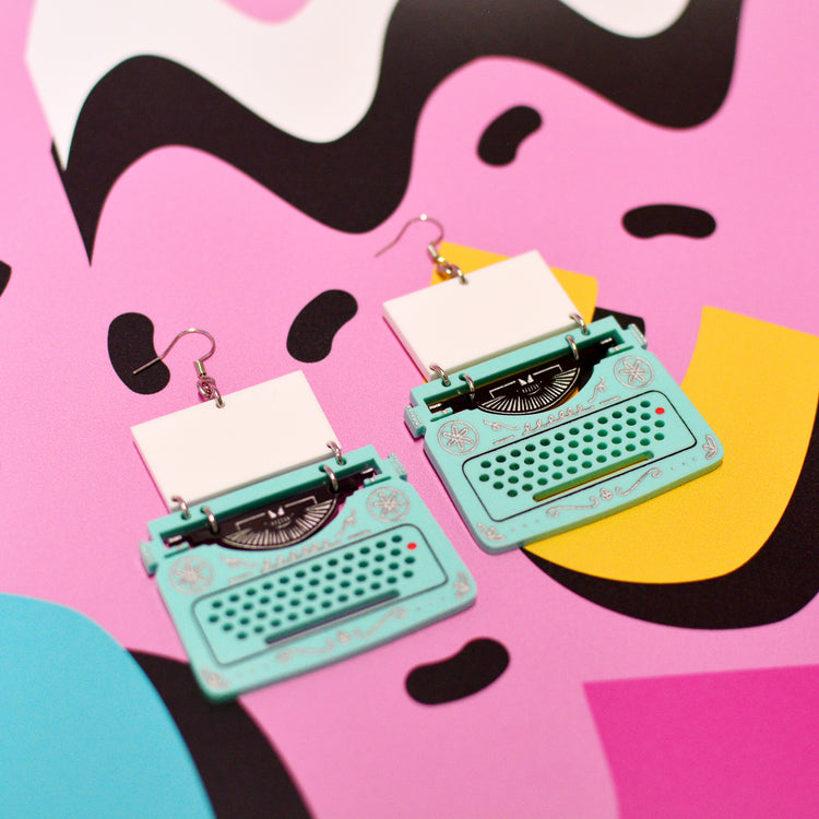 A pair of dangle earrings shaped like vintage typewriters, featuring mint green bodies, white paper stacks, and silver hooks, displayed on a vibrant pink background with abstract black and yellow shapes.