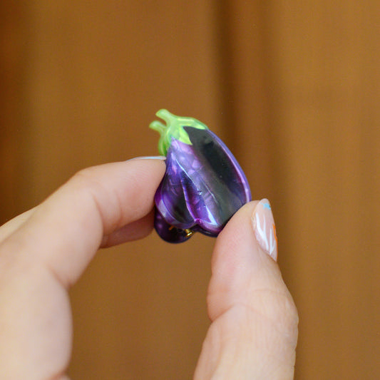 Hand holding a small purple aubergine shaped hair clip with green stem against a wooden background