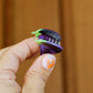 Hand holding a small purple and green aubergine shaped hair clip against a wooden background. The claw is held open so you can see the tiny transparent teeth inside.