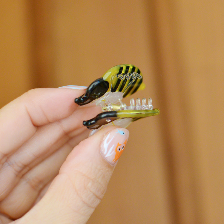 Hand holding a small bee shaped hair clip against a wood background. The claw is held open so you can see the tiny transparent teeth inside.