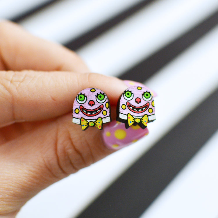 A close-up of a hand holding a pair of small, Mr Blobby stud earrings. The earrings feature a pink face with green eyes, a red nose, a wide grin, and a yellow bow tie.