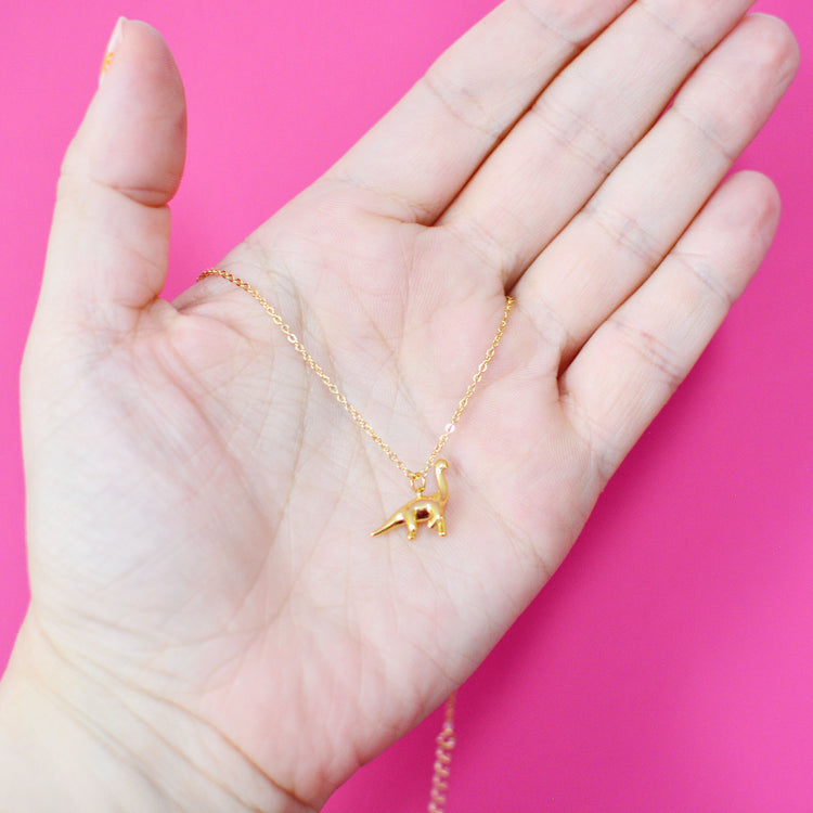 Gold necklace with a dinosaur pendant held in a hand against a pink background so you can see it is actually very small and dainty