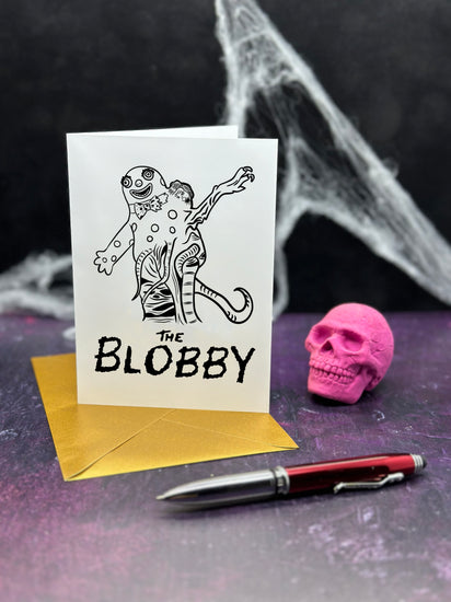 Greeting card with 'The Blobby' design (a creature that is a mash up between Mr Blobby and The Thing), gold envelope, pen, and pink skull on a dark background.