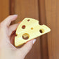 Hand holding a yellow cheese-shaped hair claw clip against a wooden background. These cheese is triangular and has holes in like emmenthal.