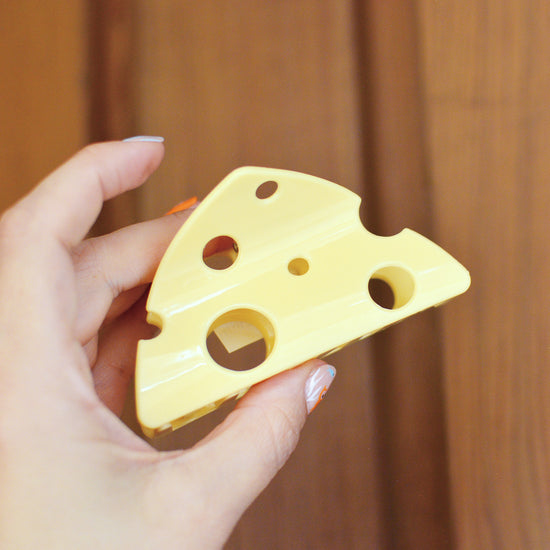 Hand holding a yellow cheese-shaped hair claw clip against a wooden background. These cheese is triangular and has holes in like emmenthal.