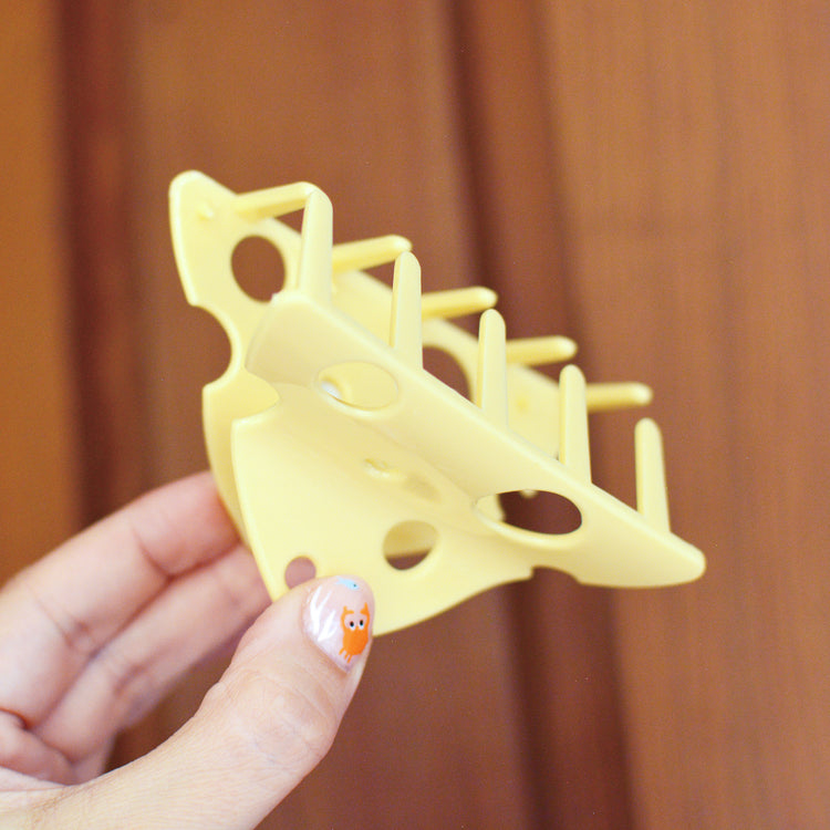 Hand holding a yellow plastic hair claw in the shape of a piece of cheese against a wooden background. The claw is held open to show the teeth that will hold your hair together.