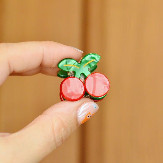 Hand holding a cherry shaped hair clip with red berries and a green stalk against a wooden background
