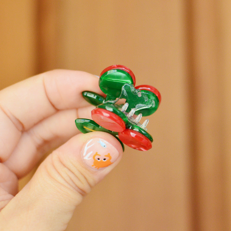Hand holding a cherry shaped hair clip with red and green berries against a wooden background. The claw is held open to reveal the transparent teeth inside.