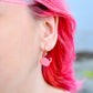 A close-up of a person's ear wearing a small pink crab-shaped dangle earring with a gold hoop against a blurred outdoor background.