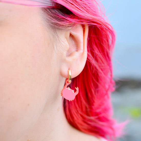 A close-up of a person's ear wearing a small pink crab-shaped dangle earring with a gold hoop against a blurred outdoor background.
