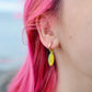 A close-up of a person's ear wearing a gold huggie hoop earring with a dangling yellow fish charm, set against bright pink hair and a blurred outdoor background.
