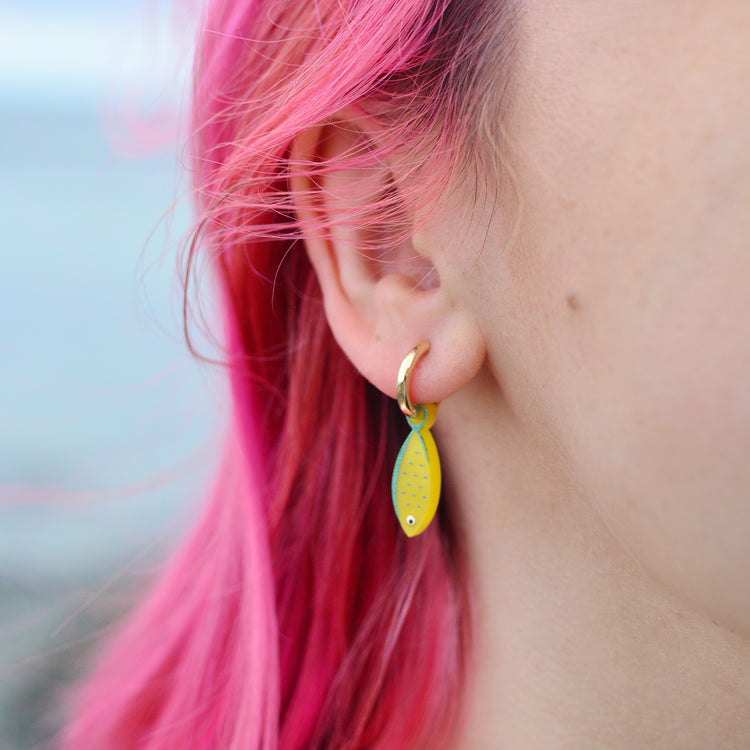 A close-up of a person's ear wearing a gold huggie hoop earring with a dangling yellow fish charm, set against bright pink hair and a blurred outdoor background.