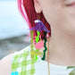 A close up of a woman's ear with her pink hair showing in the background against a blurry picture of the sea. She is wearing a statement earring of a blue wave with green seaweed hanging below. There is also a yellow fish and a coral pink crab attached to the seaweed.
