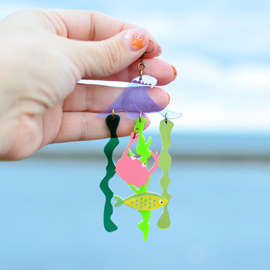 A very large earring being held up in front of the sea by a woman's hand. The earring has a sea wave shape in blue with green wiggly seaweed pieces hanging below. There is also a yellow fish and a coral pink crab hanging from the seaweed.