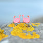 A pair of pink cartoon crab stud earrings with raised claws, resting on a gray rock covered in bright yellow lichen against a soft blue blurred background.