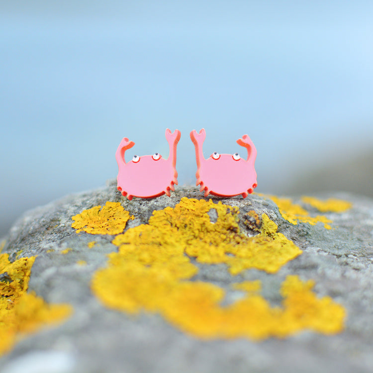 A pair of pink cartoon crab stud earrings with raised claws, resting on a gray rock covered in bright yellow lichen against a soft blue blurred background.