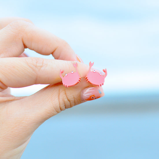 A close-up of a hand holding a pair of pink crab-shaped stud earrings against a soft, blurred blue background.