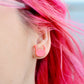 A close-up of a person's ear wearing a small pink crab stud earring, with bright pink hair visible against a softly blurred outdoor background.