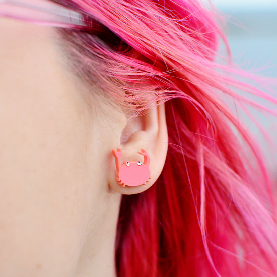 A close-up of a person's ear wearing a small pink crab stud earring, with bright pink hair visible against a softly blurred outdoor background.