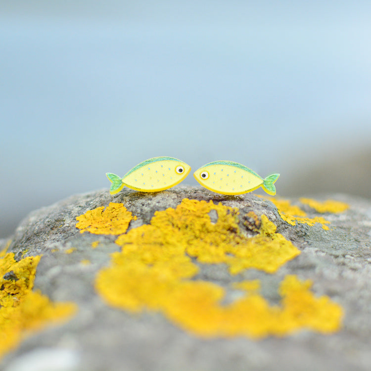 Two small fish-shaped earrings in yellow and green, resting on a rock covered in yellow lichen with a blurred blue-gray background.