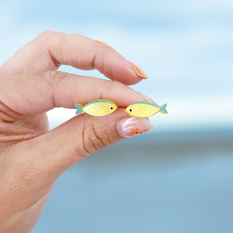 A woman hold two stud earrings between her fingers against the backdrop of the sea. The earrings are yellow fish shapes with blue details and big white eyes with black centres.