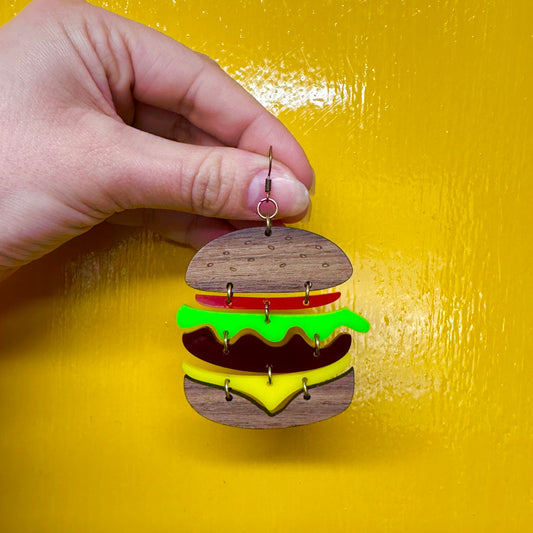 A hand holding a statement earrings designed to look like a stacked burger with wooden bun, red tomato, green lettuce, brown patty, and yellow cheese slice, against a yellow background.