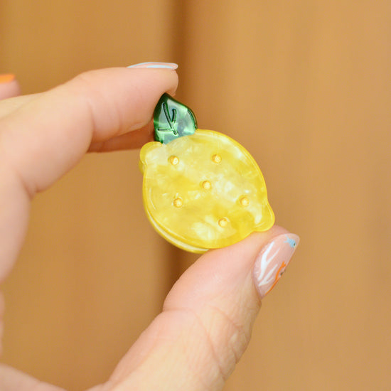 Yellow lemon-shaped hair clip with a green leaf held between fingers against a wooden background
