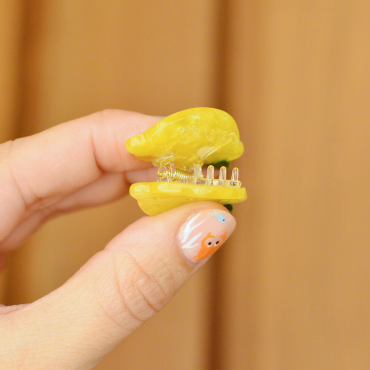 Hand holding a small yellow hair clip in the shape of a lemon against a wooden background. The claw is held open to reveal the transparent teeth inside.