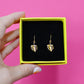 Gold plated monstera leaf earrings in a yellow box with black velvet padding, held by a hand against a pink background