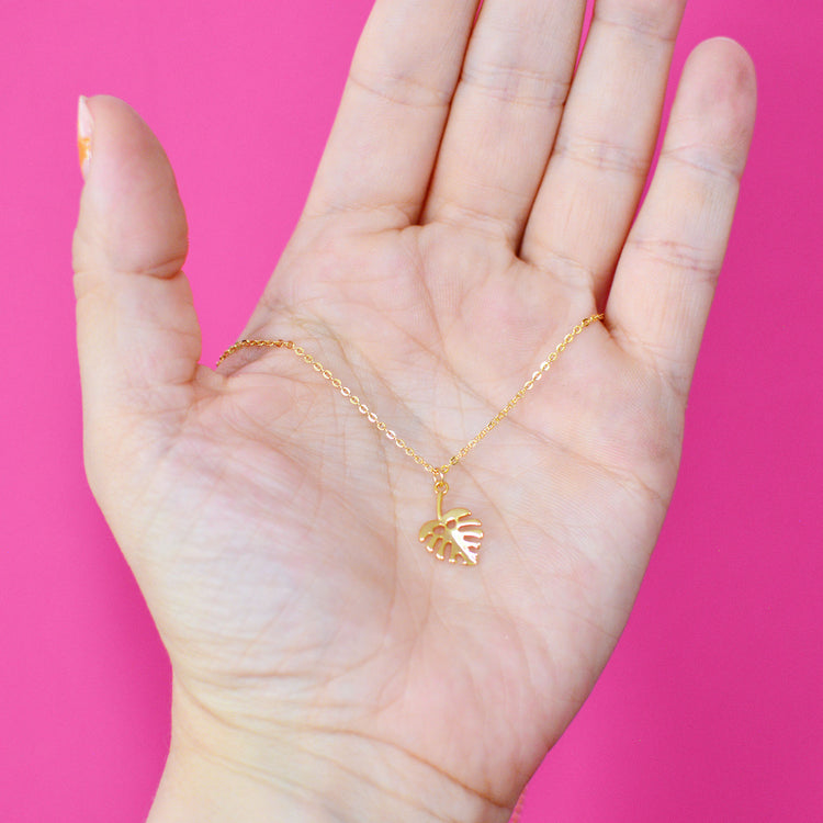 Gold plated necklace with a leaf pendant held in a hand against a pink background