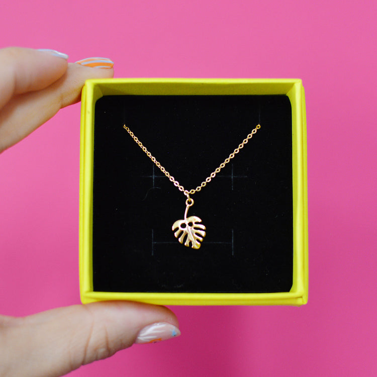 Gold plated necklace with a leaf pendant in a yellow box laid on top of a black velvet cushion against a pink background