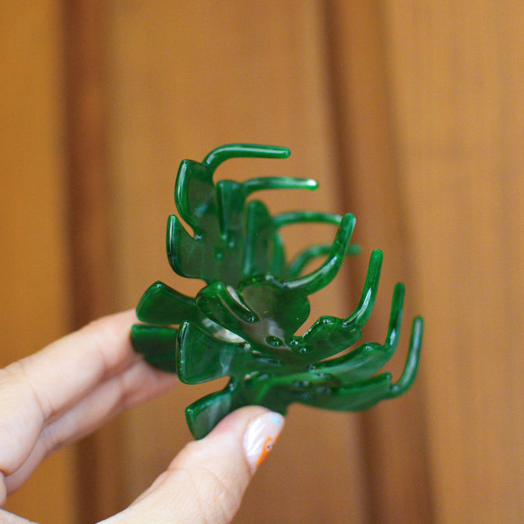 Hand holding a green plastic hair claw in the shape of a plant against a wooden background. The claw is held open to show the teeth that will hold your hair up.