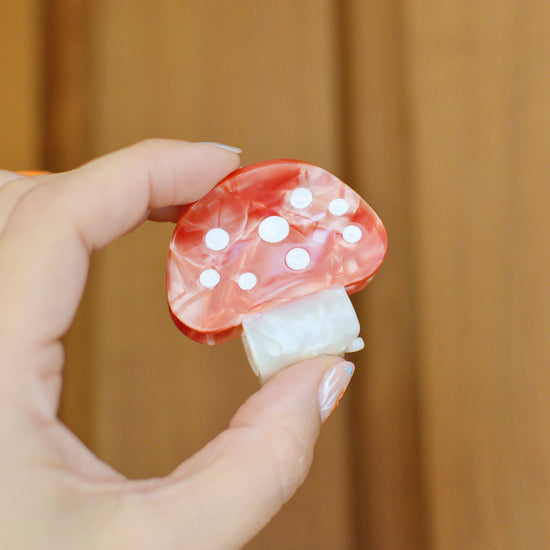 Hand holding a red and white mushroom-shaped hair claw clip against a wooden background