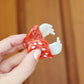Hand holding a red and white mushroom design hair claw clip against a wooden background. The claw is held open to show the teeth that will hold your hair together.