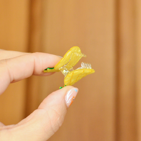 Hand holding a small yellow and green pineapple hair clip against a wooden background. The claw is held open so you can see the transparent teeth inside.