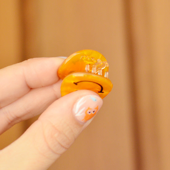 Hand holding a yellow smiley face hair clip against a wooden background. The claw is held open so you can see the transparent teeth inside.