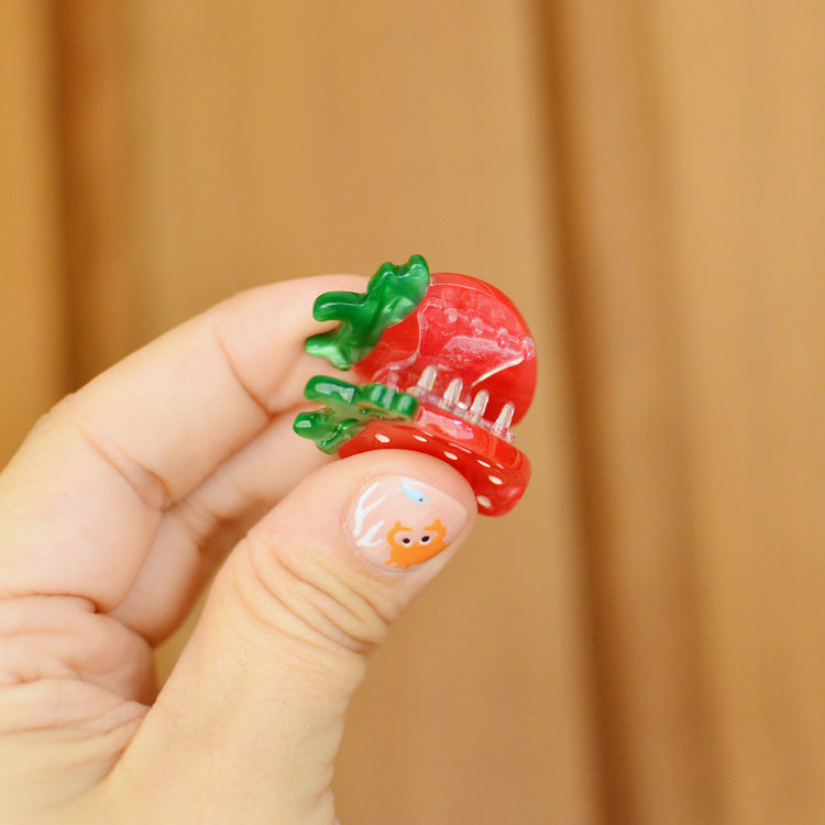 Hand holding a small red and green strawberry hair clip against a wooden background. Claw is held open to reveal the transparent teeth inside.