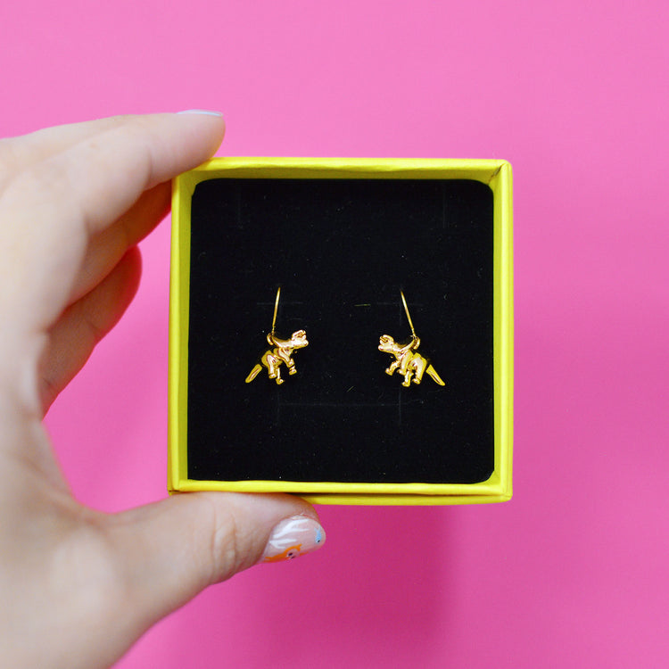 Gold triceratops dinosaur shaped earrings displayed in a yellow box with black velvet cushion, held by a hand against a pink background